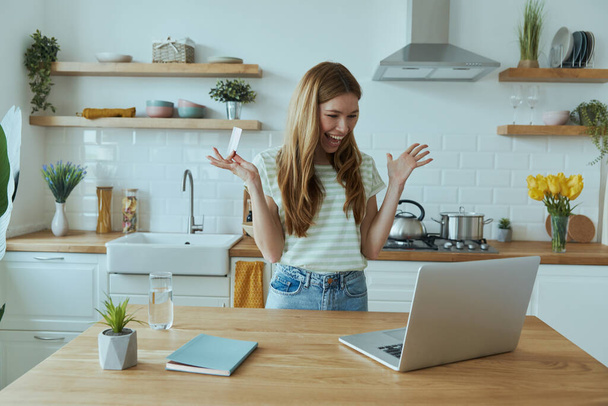 Excited young woman enjoying online shopping and using credit card while sitting at the kitchen - Фото, зображення
