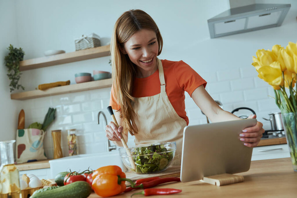 Впевнена молода жінка готує і використовує цифровий планшет, стоячи на домашній кухні
 - Фото, зображення