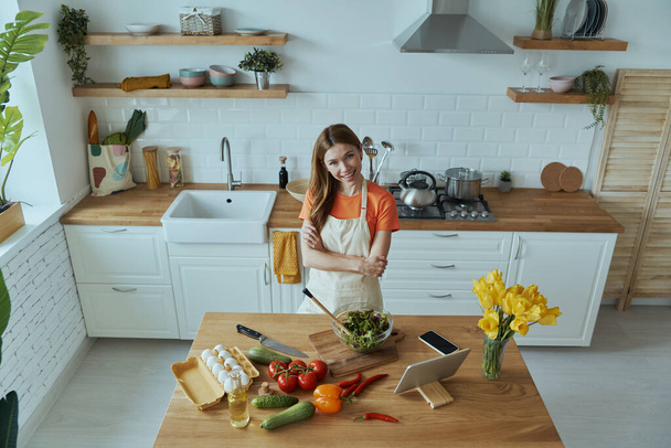 Top view of beautiful young woman looking at camera and smiling while cooking at the kitchen - Foto, afbeelding