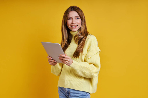 Happy young woman holding digital tablet while standing against yellow background - Фото, изображение
