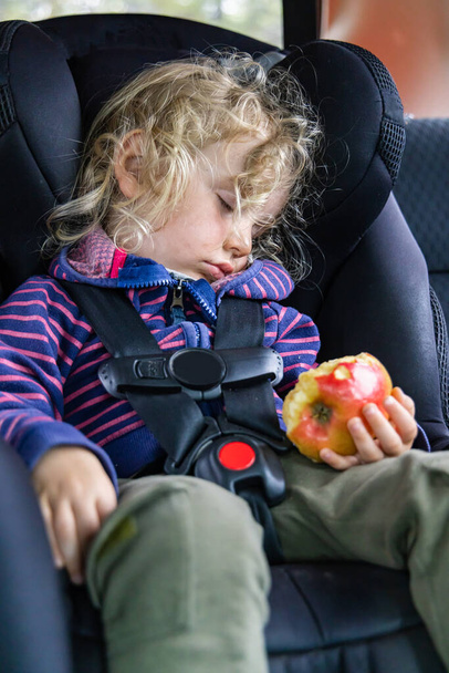 Ein entzückender dreijähriger Junge schläft in einem Kindersitz auf der Rückbank eines Autos während einer Autofahrt und schläft ein, während er an Apfel nascht. - Foto, Bild