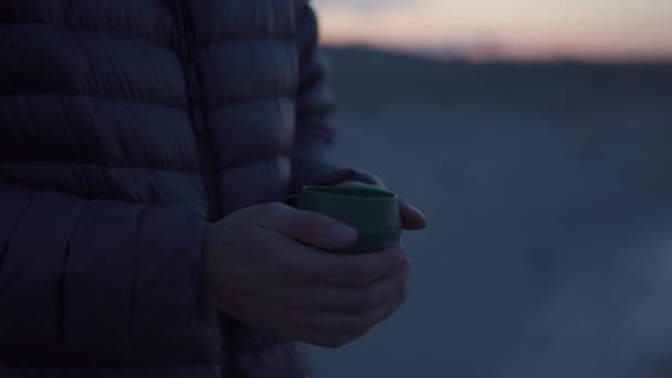 ハイキングキッチン。ダウンジャケットの男の旅行者の手が凍り、日没のキャンプ場で熱いキャンプカップに彼の腕を暖めます。折り畳み式のカップの中で熱い飲み物.コーヒーを屋外で。登山家がお茶を飲む - 映像、動画