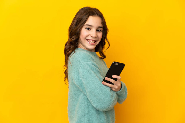 Menina caucasiana pouco isolado no fundo amarelo segurando um telefone celular e com os braços cruzados - Foto, Imagem