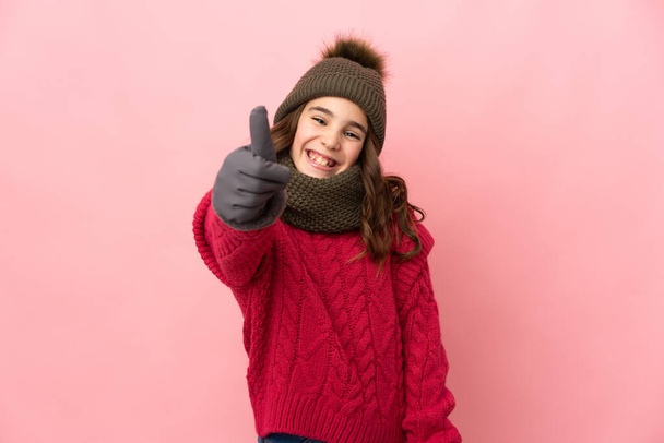Pieni tyttö talvella hattu eristetty vaaleanpunainen tausta peukalot pystyssä, koska jotain hyvää on tapahtunut - Valokuva, kuva