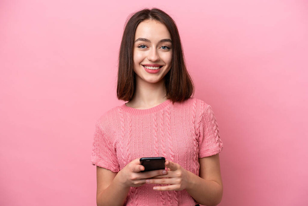 Nuori ukrainalainen nainen eristetty vaaleanpunainen tausta katselee kameraa ja hymyilee käyttäessään mobiili - Valokuva, kuva