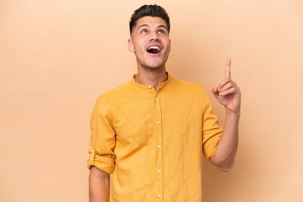 Young caucasian man isolated on beige background pointing up and surprised - Foto, imagen