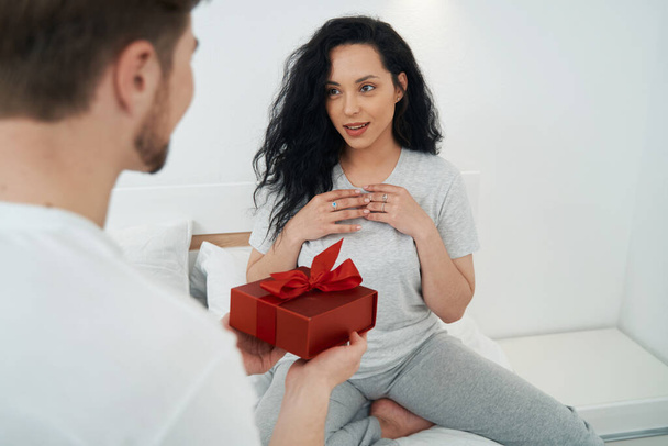 Man giving present to beloved young woman seated before him on bed - Foto, Bild