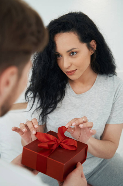 Smiling pleased lady looking at man while untying ribbon on present box - Fotografie, Obrázek