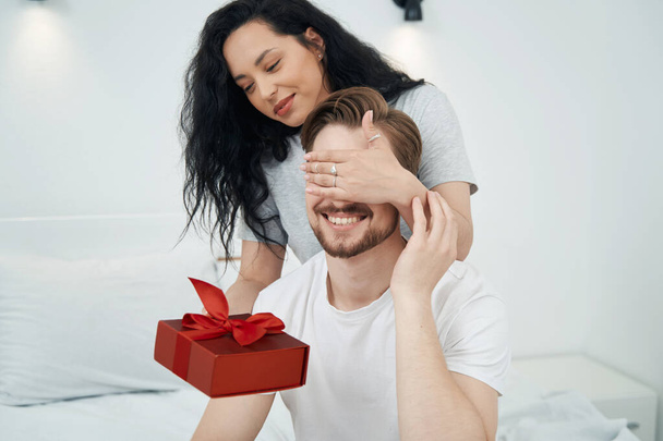 Lady giving present to smiling guy while covering his eyes with her hand - Foto, Bild
