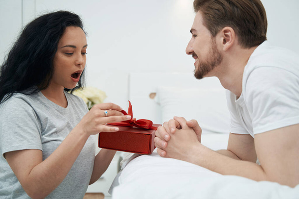 Pleased male lying on bed and watching surprised female untying ribbon on gift box - 写真・画像