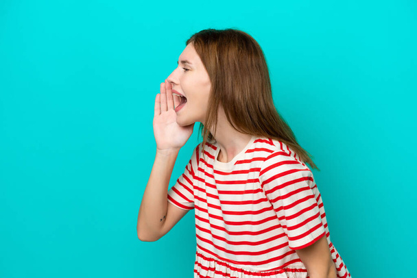 Mavi arka planda izole edilmiş genç bir İngiliz kadın ağzı sonuna kadar açık bağırıyor. - Fotoğraf, Görsel