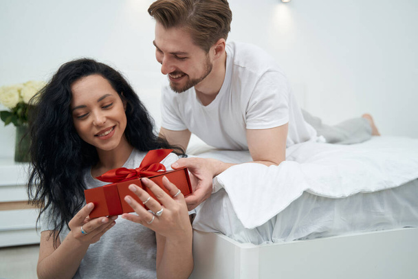 Romantic guy lying on bed and presenting gift to happy beloved lady - Foto, imagen