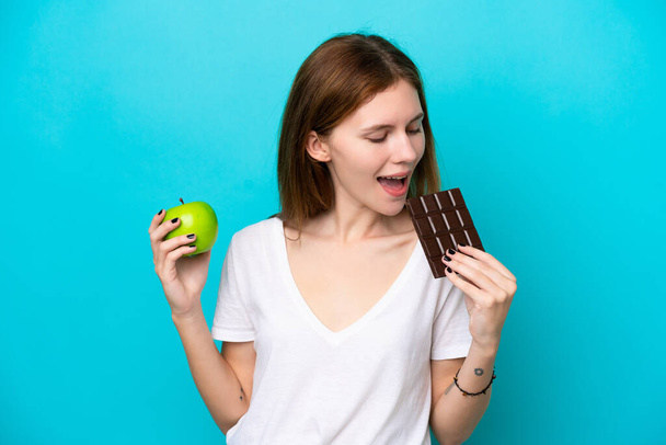 Молода англійська жінка, ізольована на синьому фоні, бере шоколадну таблетку в одній руці, а яблуко в іншій. - Фото, зображення