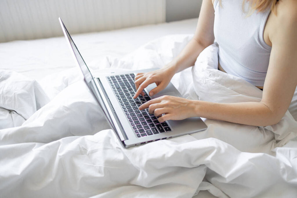家の中で午前中にベッドに座ってノートパソコンで働く幸せなカジュアルな美しい女性。家庭で働くこと、フリーランスのコンセプト. - 写真・画像