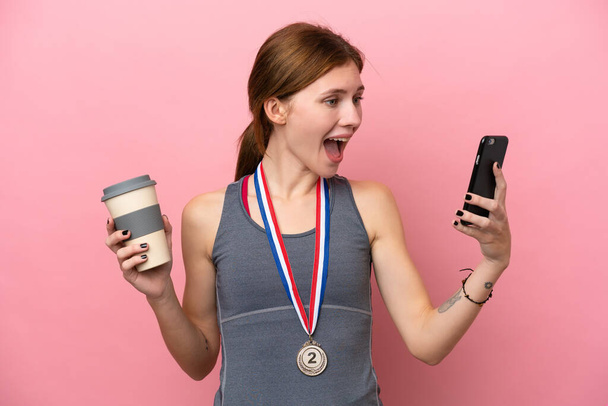 Young English woman with medals isolated on pink background holding coffee to take away and a mobile - Photo, image