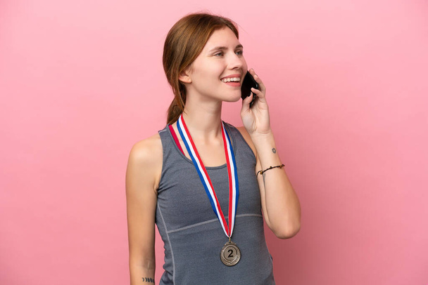 Young English woman with medals isolated on pink background keeping a conversation with the mobile phone - Photo, Image