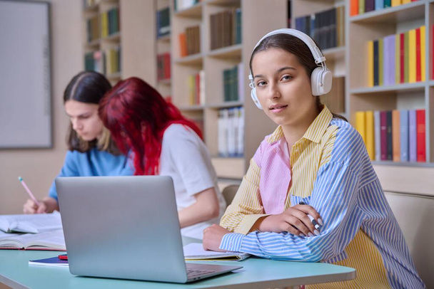 Group of teenage students study in school library classroom. In focus is teenager girl in headphones using laptop. High school, learning, education, knowledge, adolescence, people concept - Photo, Image