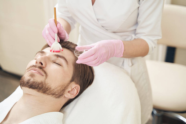 Estetista applicare liquido per peeling chimico sul viso maschile durante l'utilizzo pennello - Foto, immagini
