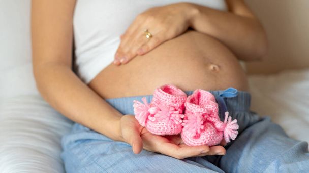 Zwangerschap baby schoenen. Gelukkige zwangere vrouw met roze babyschoenen. Kleine kinderschoen. Concept van zwangerschap, moederschap, verwachting voor de bevalling - Foto, afbeelding