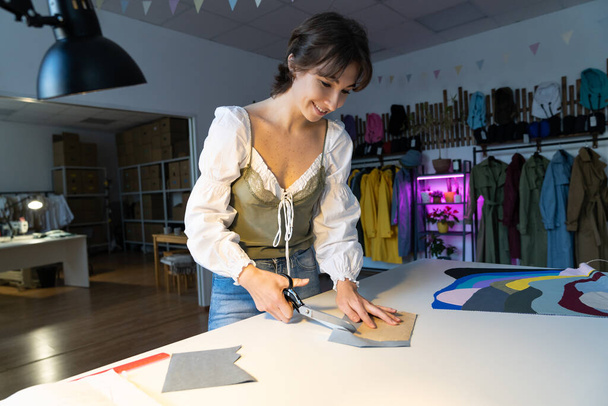 オーダーメイドの縫製ビジネスコンセプトで、若いプロのドレスメーカーはファッションコレクションをテーラリングするためのワークショップで繊維と仕事をします。ファッションデザイナーの縫製工房で織物を切断 - 写真・画像