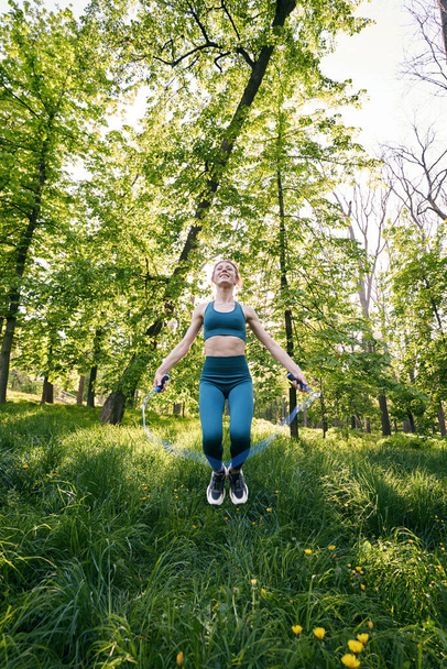 Jolly young slim female is doing workout in green wood and using jumping rope with fun - Foto, Imagen
