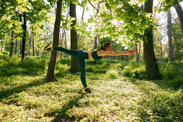 Athletic woman is standing on one foot and stretching body during yoga workout in wood - Zdjęcie, obraz
