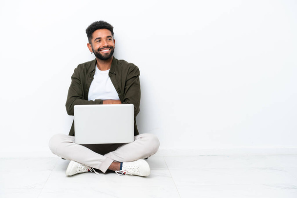 Fiatal brazil férfi laptoppal a padlón, elszigetelve fehér karokkal és boldogan. - Fotó, kép