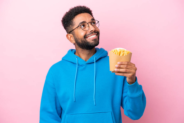 jonge Braziliaanse man met gebakken chips geïsoleerd op roze achtergrond op zoek omhoog terwijl glimlachen - Foto, afbeelding