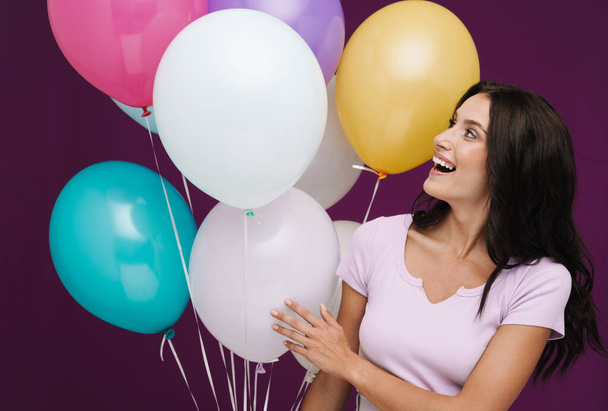 Bílá brunetka žena ve smíchu při pózování s balónky izolované přes fialové pozadí - Fotografie, Obrázek