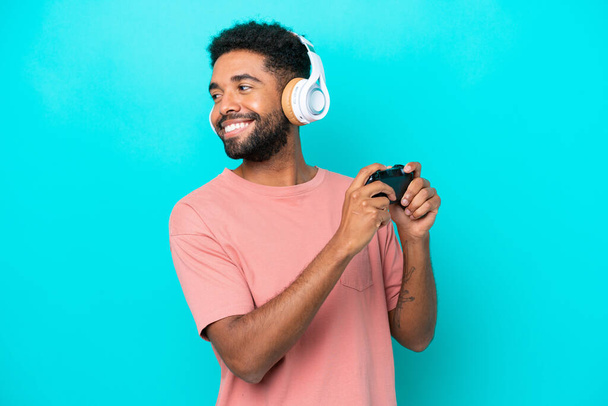 Joven brasileño jugando con un controlador de videojuegos aislado sobre fondo azul riendo en posición lateral - Foto, imagen