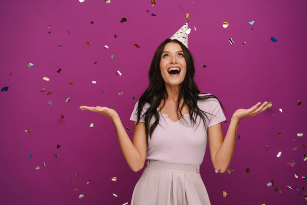 Young brunette woman in party cone making fun with confetti isolated over purple wall - Zdjęcie, obraz