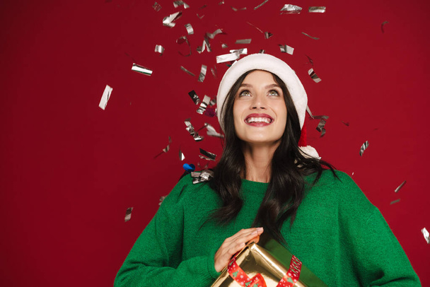 Young woman in Christmas hat posing with present box and confetti isolated over red wall - Fotografie, Obrázek