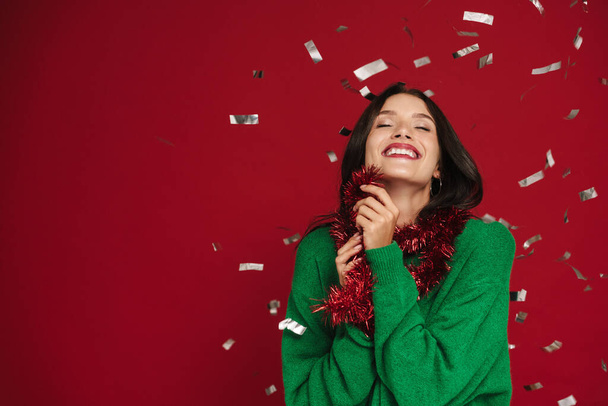 White woman laughing while posing with Christmas tinsel garland isolated over red background - Фото, изображение