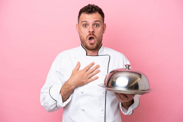 Jonge Braziliaanse chef-kok met dienblad geïsoleerd op roze achtergrond verrast en geschokt terwijl rechts kijken - Foto, afbeelding