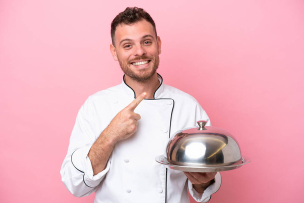 Молодий бразильський шеф - кухар з тацею, ізольованим на рожевому фоні, який вказує на бік, щоб представити продукт. - Фото, зображення