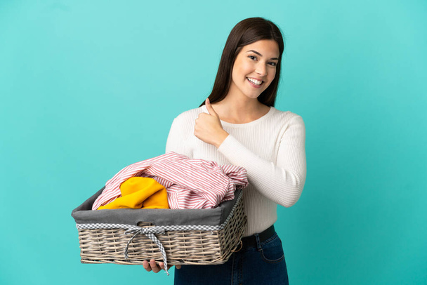 Teenager Brazilian girl holding a clothes basket isolated on blue background giving a thumbs up gesture - Foto, Imagen