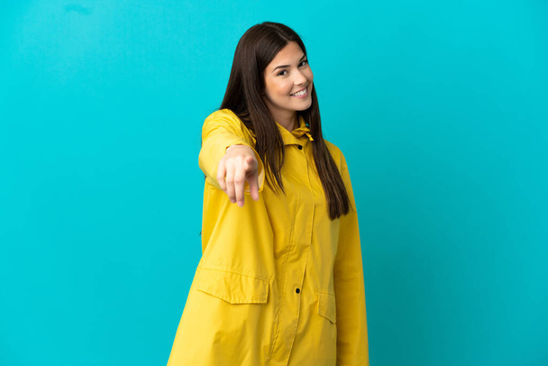 Teenager Brazilian girl wearing a rainproof coat over isolated blue background points finger at you with a confident expression - Foto, Bild