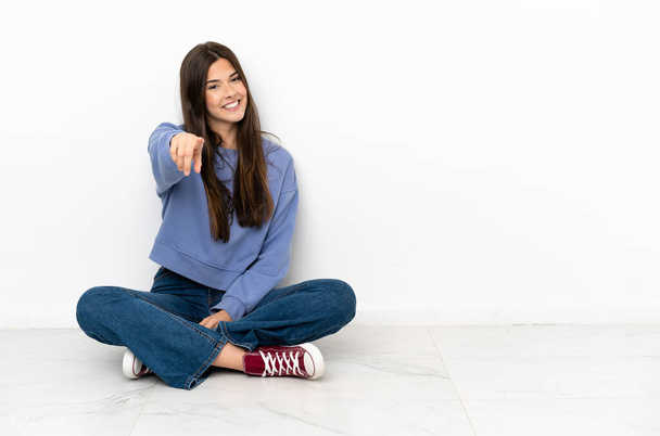 Jovem mulher sentada no chão aponta o dedo para você com uma expressão confiante
 - Foto, Imagem