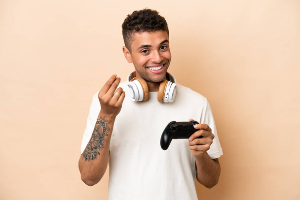 Молодий бразільський чоловік грає з відеоігровим контролером ізольованим на бежевому фоні, заробляючи гроші жестом - Фото, зображення