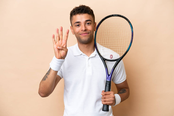młody brazylijski przystojny mężczyzna gra w tenisa na beżowym tle szczęśliwy i liczyć cztery palce - Zdjęcie, obraz