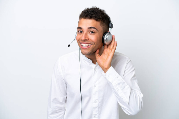 Hombre brasileño telemarketer trabajando con un auricular aislado sobre fondo blanco escuchando algo poniendo la mano en la oreja - Foto, Imagen