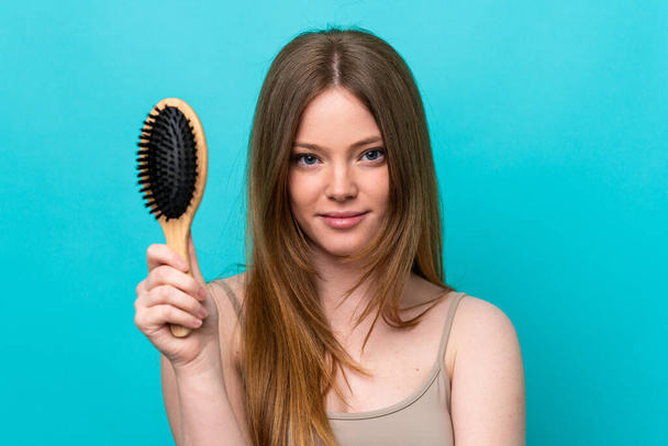 Young caucasian woman isolated on blue background with hair comb - Foto, afbeelding