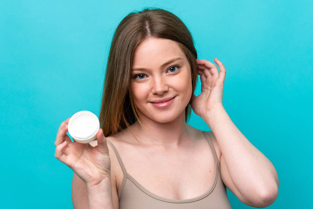Young caucasian woman isolated on blue background with moisturizer and happiness - Foto, Imagen