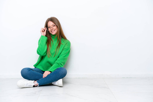 Young caucasian woman sitting on the floor isolated on white background with glasses and happy - 写真・画像