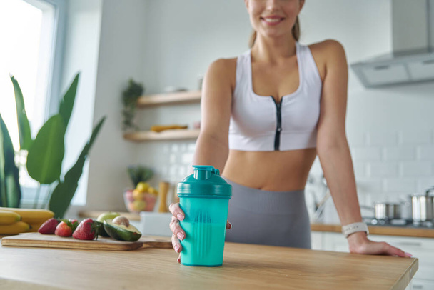 Close-up of woman in sports clothing holding shaker with protein cocktail at the kitchen - Foto, afbeelding