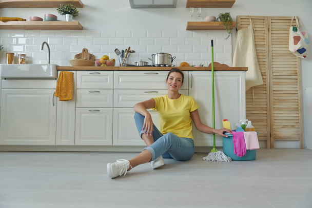Cheerful young woman sitting on the floor near the bucket with cleaning stuff at the kitchen - Zdjęcie, obraz