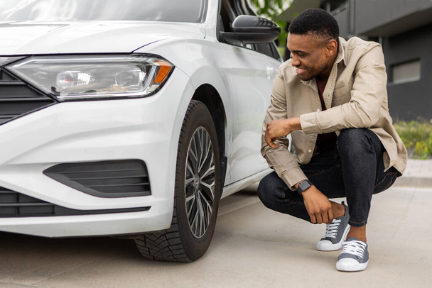 Ein junger schwarzer Mann sitzt neben dem Auto und schaut auf das Lenkrad, das die Reifen eines neuen Autos auf eine neue Felge prüft. Hochwertiges Foto - Foto, Bild