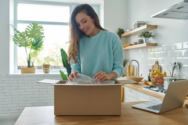 Attractive young woman unpacking box while standing at the domestic kitchen - Foto, imagen