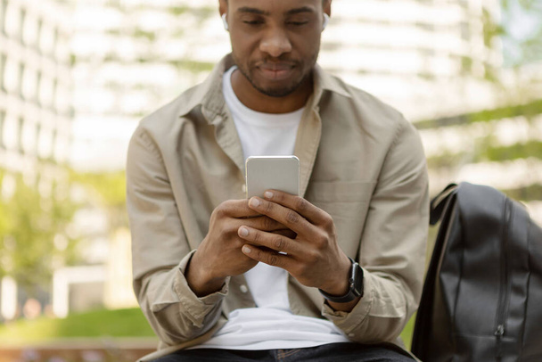 A black guy in headphones sits on a bench in the city with a smartphone. High quality photo - Fotoğraf, Görsel