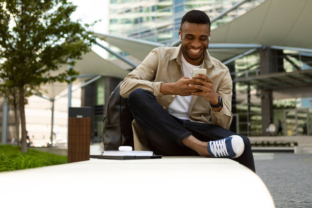 Африканський американець, який користується смартфоном, сидить на лавці в місті з навушниками. Фотографія високої якості - Фото, зображення